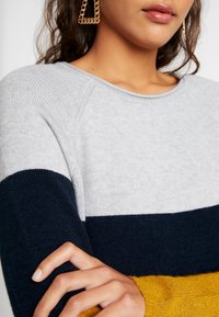 Maglione a blocchi di colore con sezioni grigie, blu navy e senape. Tessuto morbido, scollatura tonda e dettagli a coste sulle maniche.