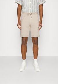 Beige cotton shorts with an elastic waistband and drawstring, featuring rolled cuffs. Paired with white sneakers and a striped shirt.