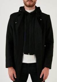 Abrigo negro con capucha y cierre de cremallera, presenta un diseño de doble cremallera, bolsillos laterales y una textura suave. Lleva encima un suéter blanco de canalé.