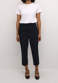 T-shirt en coton blanc associé à un pantalon cropped bleu marine foncé, à taille haute et texture lisse, complété par des talons en cuir verni noir.