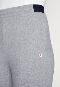 Pantaloni della tuta grigi con una vita blu navy, dotati di un piccolo logo sul lato. Il materiale sembra morbido con una finitura testurizzata.