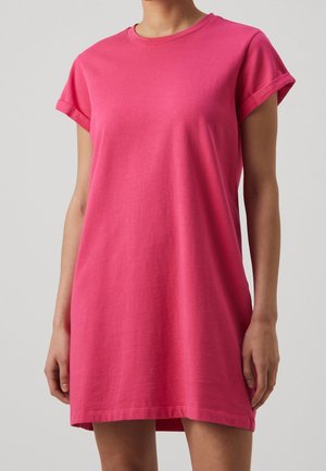 Mujer con vestido informal de manga corta de color rosa brillante con las mangas remangadas, de pie frente a un fondo liso de color gris claro.