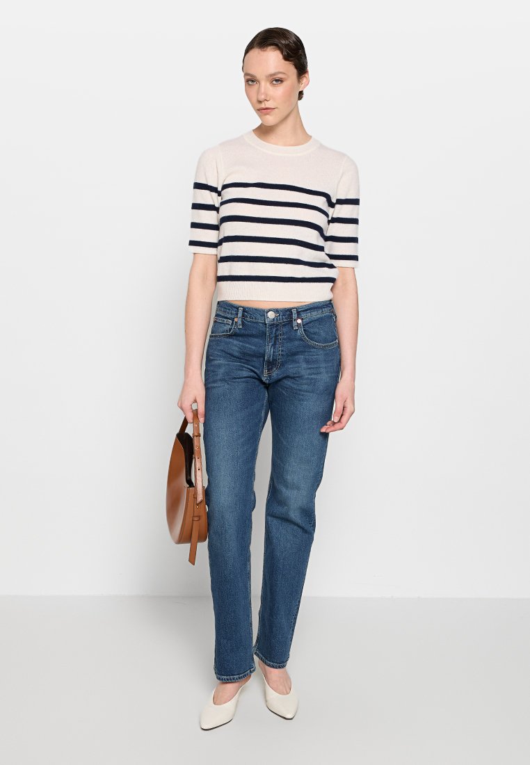 Gestreifter Strickpullover in Creme und Marineblau, cropped Design. Blaue Jeans, gerader Schnitt, mit leichter Ausbleichung. Modell hält eine braune Tasche und trägt beige Absätze.