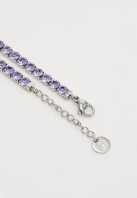 Violetinis kristalų apyrankė su sidabro spalvos metalo detalėmis, turinti omaro tipo užsegimą ir mažą logotipo žymę. Tekstūruoti grandinėlės jungia kiekvieną kristalą.