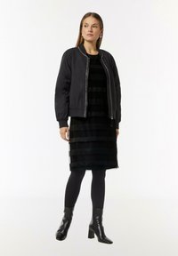 Schwarze Bomberjacke mit gerippten Bündchen, kombiniert mit einem schwarz gestreiften Kleid. Das Outfit umfasst schwarze Strumpfhosen und klobige Stiefel mit Absatz.