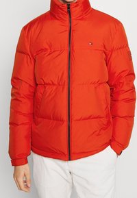 Man som bär en livlig röd vadderad jacka med framdragkedja och två fickor, tillsammans med vita byxor, stående mot en enfärgad bakgrund.