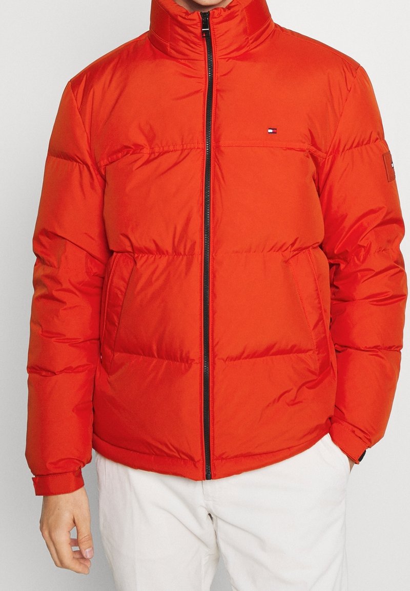 Un homme portant une veste matelassée rouge vif avec fermeture éclair à l'avant et deux poches, assortie à un pantalon blanc, se tenant devant un fond uni.