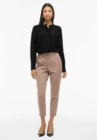 Melnā blūze ar garām piedurknēm, ar ruffle apkakli un pogu detaļām, saskaņota ar bēšiem piegriezuma buksēm un melniem kurpēm ar norādītu purngalu.