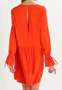 Pitkähihaista oranssia mekkoa, jossa on aukko selässä, kerätty vyötärö ja sidottavat hihansuut. Pehmeä kangas, jossa on rento, virtaava siluetti.