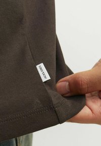 Camiseta de algod�ón marrón mostrando una mano sosteniendo el dobladillo y una pequeña etiqueta blanca con "JACK & JONES" impresa. Textura suave, diseño ajustado.