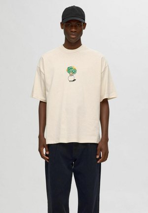 Beige bomulds oversized t-shirt med et farverigt, potteret plantegrafik på brystet. Sammen med mørke bukser og en sort kasket.