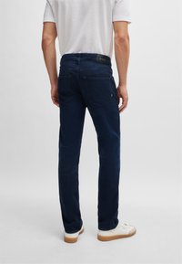 Jean en denim bleu foncé avec une coupe droite et deux poches arrière. Une étiquette en cuir noir avec le logo de la marque sur la ceinture. Baskets blanches décontractées.