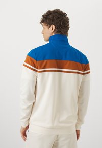 Chaqueta ligera con cremallera, que presenta un patrón de bloques de color azul, marrón y blanco, con un alto cuello y puños acanalados. Tejido texturizado.
