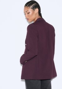 Bordeauxroter Blazer mit strukturiertem Design, ausgestattet mit goldenen Knöpfen, tailliertem Schnitt und glatter Textur. Von hinten dargestellt.