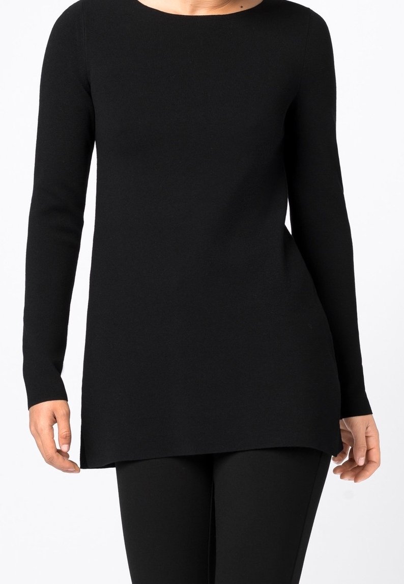 Femme portant un haut tunique noir à manches longues associé à un pantalon noir, montrée du cou à mi-cuisse sur fond blanc.