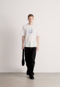 Camiseta blanca de manga corta con gráfico de una carita sonriente azul, combinada con pantalones cargo negros holgados y un bolso de hombro negro. Fondo neutro.