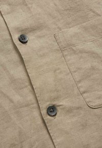 Beige linnedugsskjorteafsnit med mørke runde knapper og en syet lomme på højre side af beklædningsgenstanden.