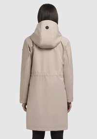 Manteau isolé beige avec capuche, taille cintrée et manches longues. Présente un accent logo rond au dos de la capuche. Texture lisse.