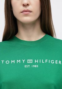 Πράσινο βαμβακερό t-shirt με στρογγυλή λαιμόκοψη, με λευκό κείμενο: "TOMMY HILFIGER EST. 1985," τοποθετημένο κεντρικά στο στήθος.