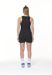 Crni majica bez rukava u kombinaciji s crnim sportskim šortsevima. Model nosi bijele i plave tenisice s bijelim čarapama na kojima je crni natpis.