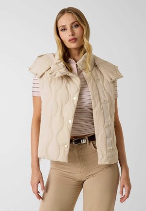 Frau trägt beige gesteppte ärmellose Weste über rosa gestreiftem Poloshirt und beiger Hose, steht vor einfachem weißem Hintergrund.