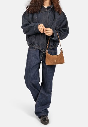 Vrouw die een oversized donkere denimjas en broek draagt, met een kleine bruine suède handtas met versierde sluiting in haar hand.