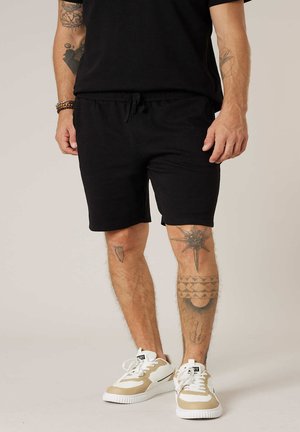 Homme portant un short noir et un t-shirt noir, des baskets beige et blanches, avec des tatouages visibles sur les bras et les jambes.