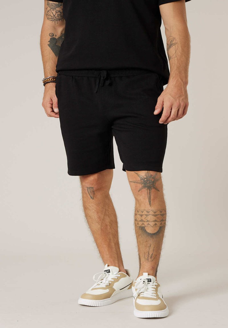 Homme portant un short noir et un t-shirt noir, des baskets beige et blanches, avec des tatouages visibles sur les bras et les jambes.