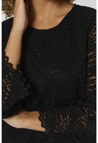 Femme portant un haut noir à manches longues en dentelle à motifs floraux avec des bords festonnés, touchant son cou d'une main tandis que l'autre traverse son corps.