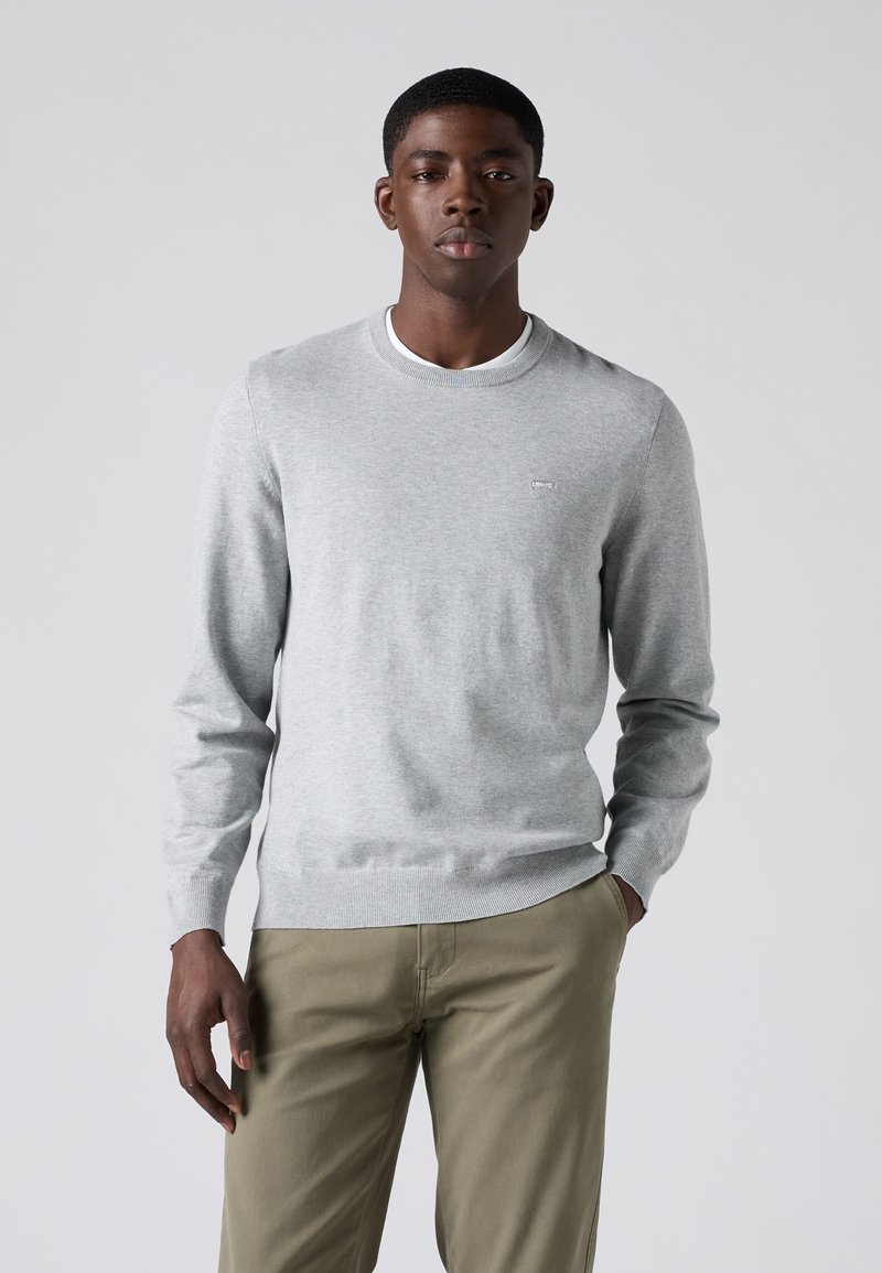 Jonge man die een lichtgrijze trui met ronde hals en beige broek draagt, staat met één hand in de zak tegen een effen achtergrond.