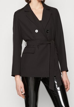 Nainen, joka pukeutunut mustaan vyölliseen bleiseriin, jossa on kaksi eriväristä nappia, ja kiiltäviin mustiin vinyylihousuihin, seisoo tasaisen taustan edessä.