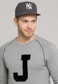 Sudadera gris con una gran "J" negra en el frente, combinada con una gorra de béisbol gris oscuro con el logo blanco de los New York Yankees.