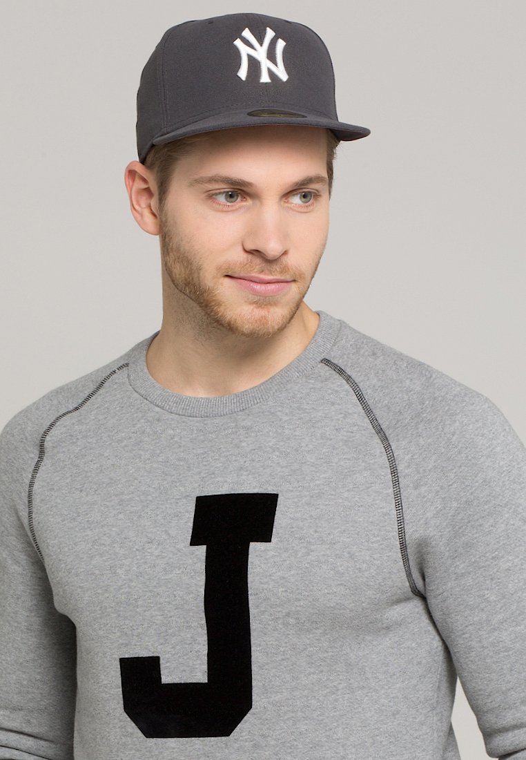 Sudadera gris con una gran "J" negra en el frente, combinada con una gorra de béisbol gris oscuro con el logo blanco de los New York Yankees.