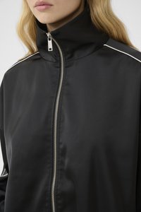 Chaqueta negra con cremallera, hecha de una tela suave y ligera. Presenta un cuello alto, cremallera plateada y franjas de acento blancas en los hombros.