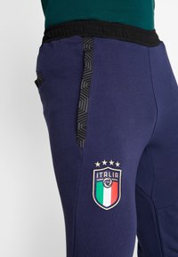 Marineblauwe sweatpants met een zwarte elastische tailleband en een geometrisch patroon aan de zijkant; voorzien van het logo van het nationale voetbalteam van Italië op de dij.