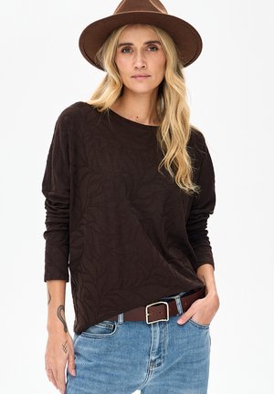 Brązowa koszula z długim rękawem w liściasty wzór, miękka w dotyku, o luźnym kroju, zestawiona z niebieskimi dżinsami i brązowym paskiem.