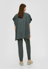 Overmaatse, donkergroene top met korte mouwen, gladde textuur, gecombineerd met bijpassende slim-fit broeken en lichtgekleurde sneakers.