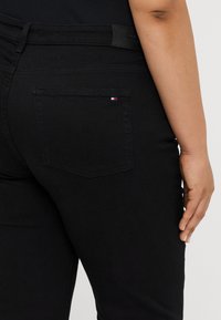 Jean en denim noir avec une texture lisse, présentant une poche arrière avec un petit patch de logo rouge, blanc et bleu et une taille texturée.