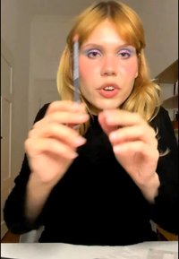 Junge Frau mit blondem Haar und lila Lidschatten hält einen Stift und spricht direkt in die Kamera in einem Innenraum.