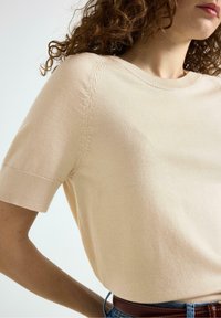 Maglietta a maniche corte lavorata a maglia color beige chiaro con scollo rotondo, dettagli a coste sulle maniche e una consistenza liscia. Indossata con jeans in denim blu.