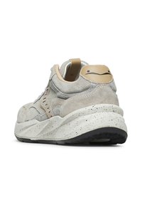 Sneaker in suede beige con suola bianca maculata, suola nera e una linguetta sul tallone color cuoio con un design di volto minimalista, mostrato da un angolo posteriore.