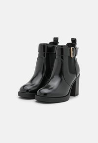 Par de botas negras brillantes hasta el tobillo con tacones de bloque, paneles elásticos laterales y correas con hebillas doradas sobre un fondo blanco liso.