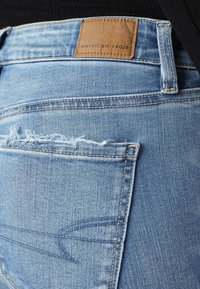 Svetle modré džínové kalhoty s roztrhanou zadní kapsou a béžovou koženou nášivkou s embosovaným nápisem "American Eagle" na pasu.