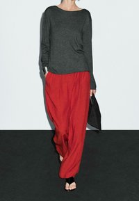 Grå langærmet top, røde løstsiddende bukser med lommer, sorte flade sandaler og en lille sort taske holdt i hånden. Teksturerede stoffer.