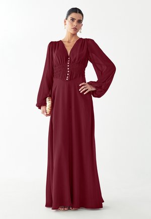 Frau steht mit der Hand in der Hüfte, trägt ein langes burgunderrotes Kleid mit Puffärmeln und hält eine goldene Clutch.