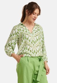 Green patterned blouse with a v-neck and long sleeves, featuring varying shades of green leaf-like shapes, paired with green trousers.