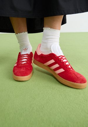Osoba nosząca czerwone buty sportowe Adidas z różowymi paskami i białe skarpetki stoi na jasnozielonym dywanie.