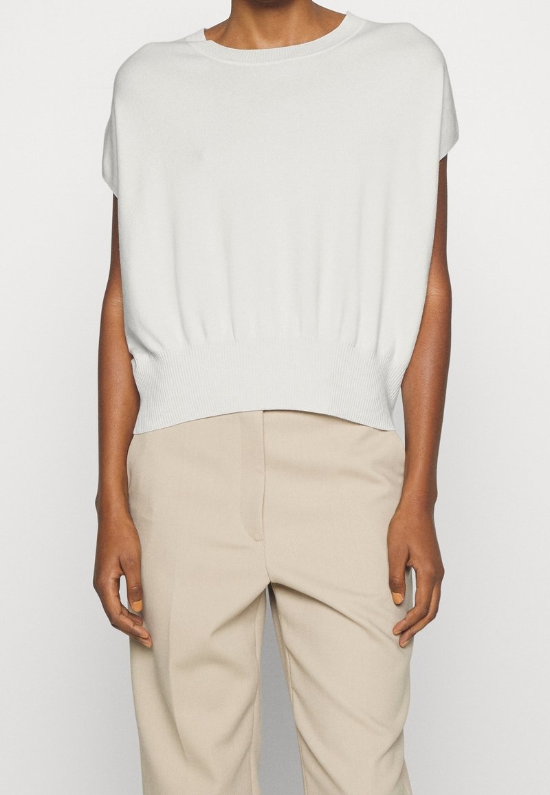 Bílý pletený top s krátkými rukávy a nařaseným pasem. Hladká textura a kulatý výstřih, v kombinaci s světle béžovými kalhotami.