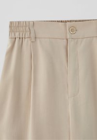 Beige broek met knoopsluiting, riemlussen en een elastische tailleband voor aanpasbare pasvorm, weergegeven op een effen lichte achtergrond.
