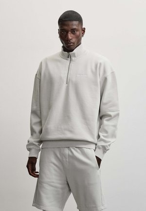 Ung mand med kort hår iført lysegrå lynlås-sweatshirt og matchende shorts, stående mod en ensfarvet lys baggrund.